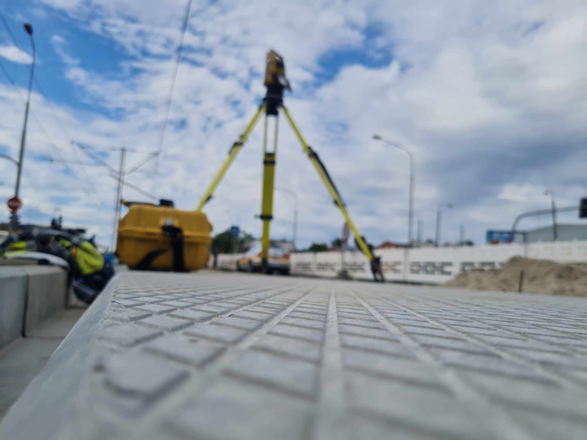 Prace pomiarowe na torowisku: żółty teodolit na trójnogu ustawiony na nowej, szarej nawierzchni, w tle błękitne niebo z obłokami.