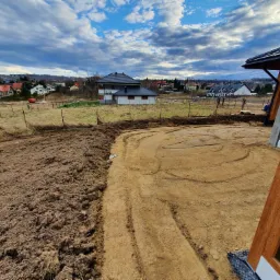 Teren po wyrównaniu, przygotowany pod budowę, widoczne ślady opon, w tle zabudowa jednorodzinna i pochmurne niebo.