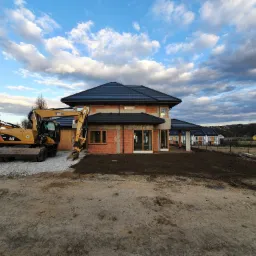 Żółta koparka CAT obok domu w trakcie budowy, widoczne ściany z czerwonej cegły, czarny dach i przygotowany teren pod ogród, w tle zabudowa jednorodzinna i zachmurzone niebo.