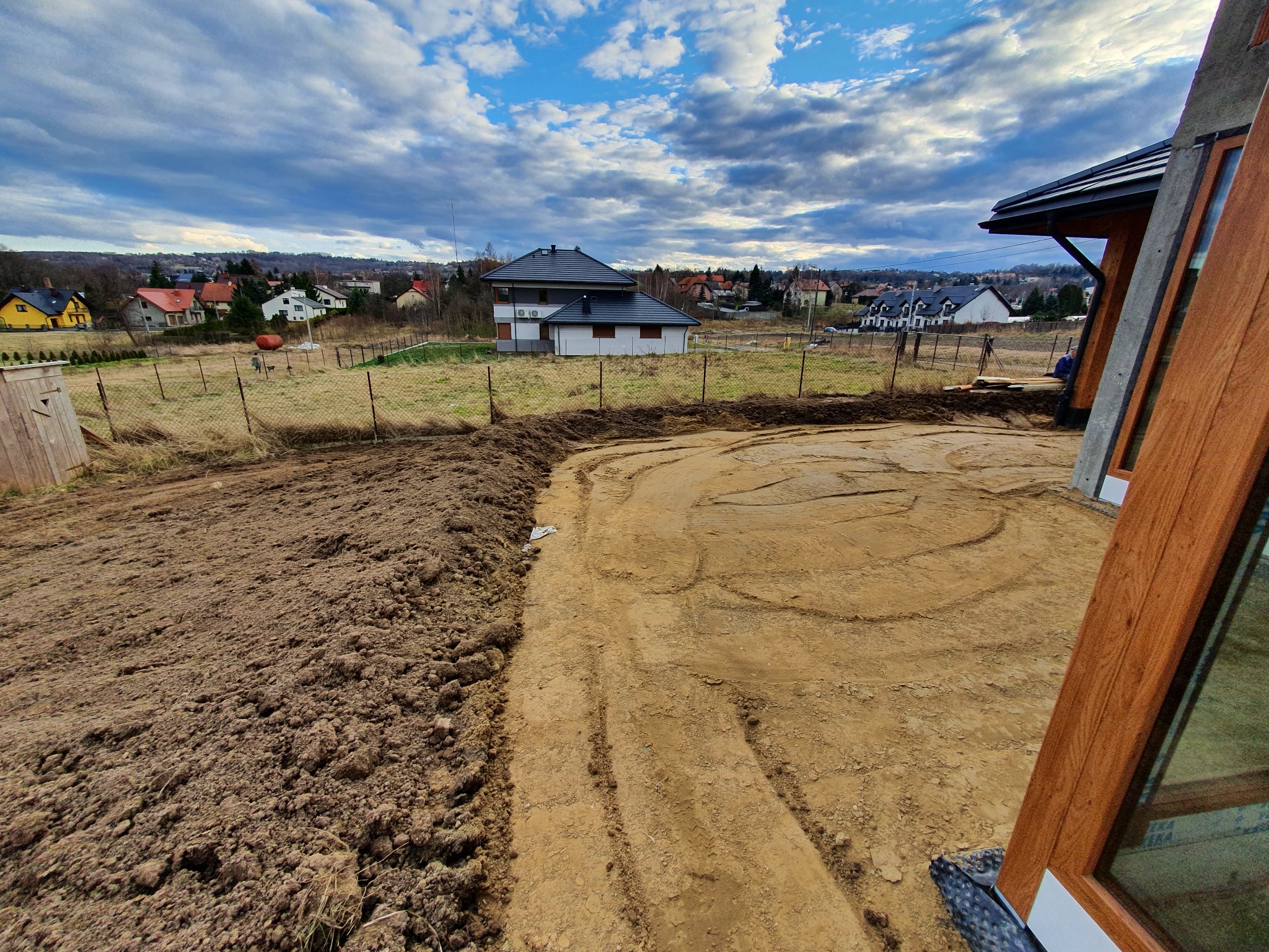 Teren po wyrównaniu, przygotowany pod budowę, widoczne ślady opon, w tle zabudowa jednorodzinna i pochmurne niebo.