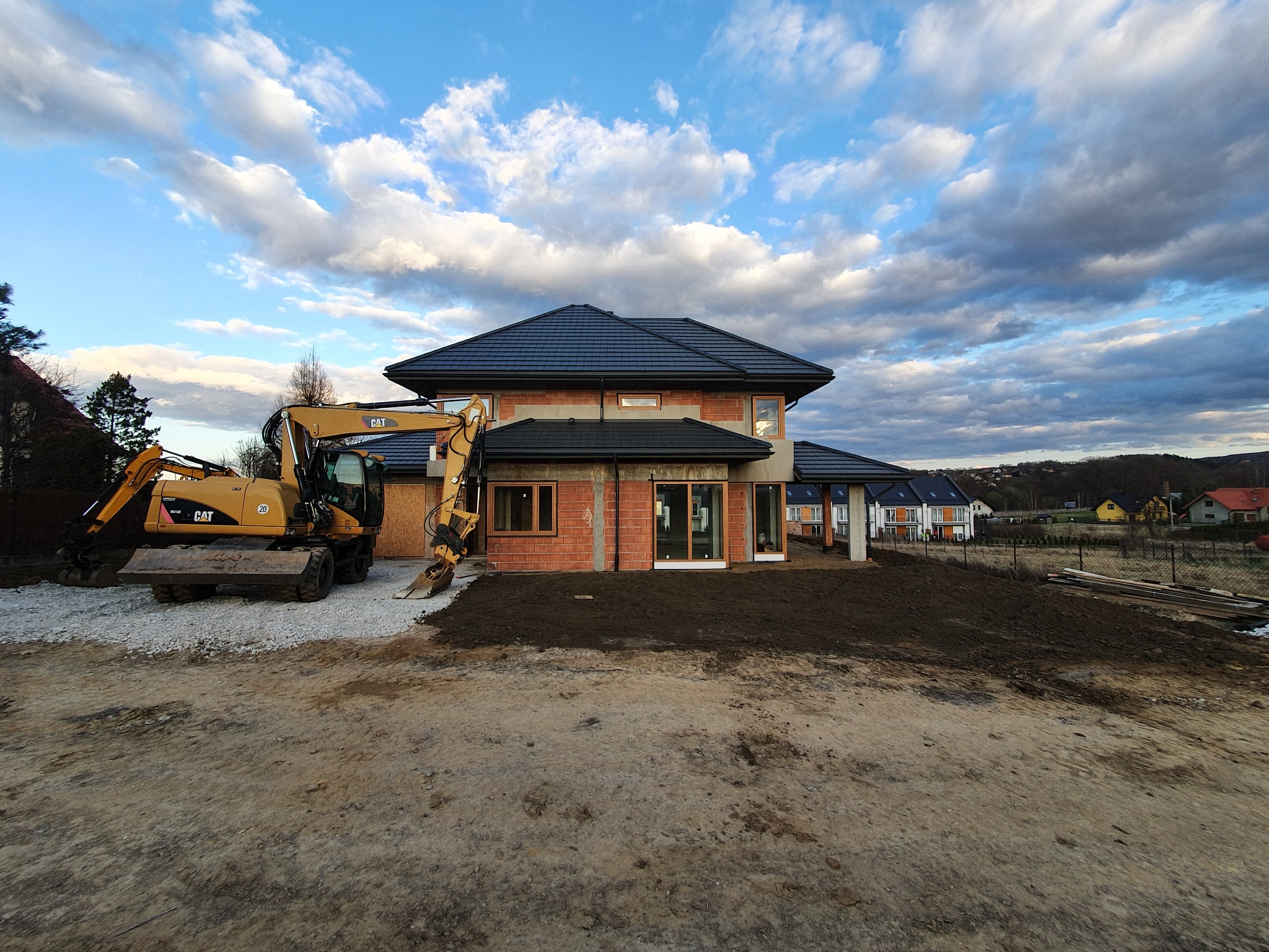 Żółta koparka CAT obok domu w trakcie budowy, widoczne ściany z czerwonej cegły, czarny dach i przygotowany teren pod ogród, w tle zabudowa jednorodzinna i zachmurzone niebo.