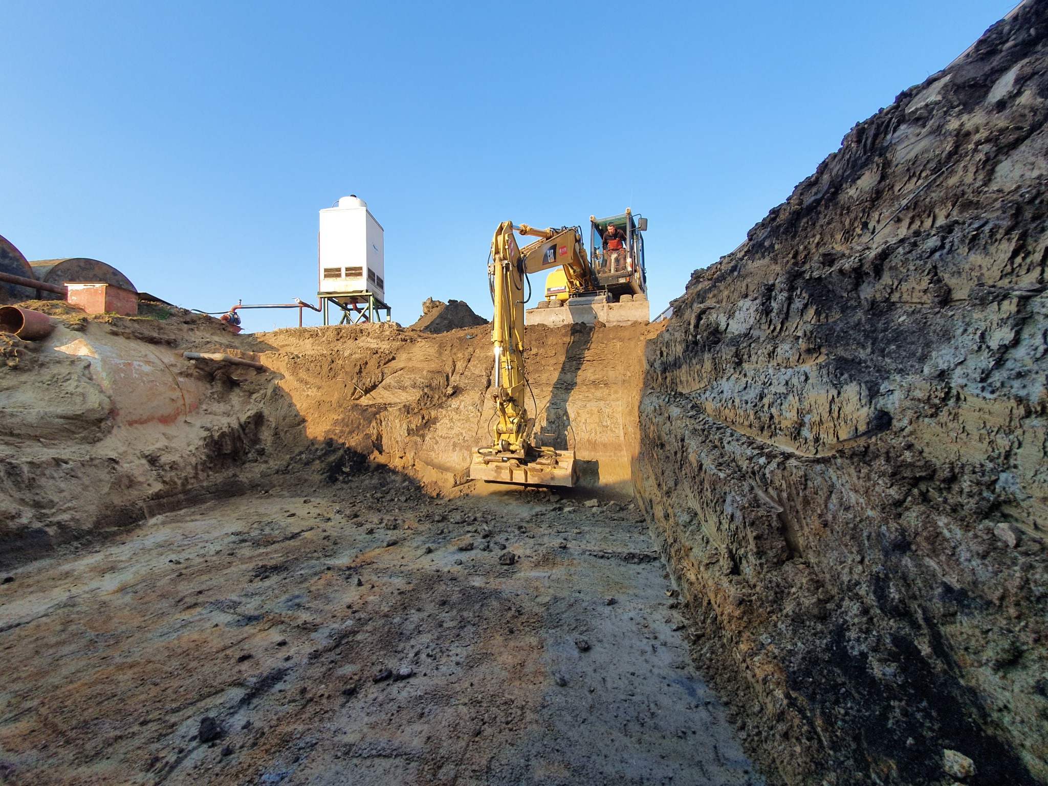 Żółta koparka w wykopie, widoczne warstwy ziemi i maszyny w tle pod błękitnym niebem.