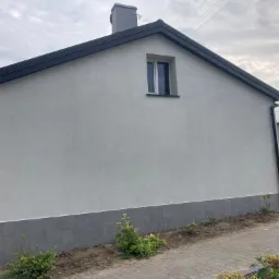 Świeżo otynkowana ściana domu jednorodzinnego z szarą podmurówką i ciemnoszarą blachodachówką, widoczne rynny i małe okno na poddaszu, w tle pochmurne niebo.