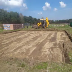 Żółta koparko-ładowarka przygotowuje teren pod budowę, widoczne wykopane obrysy fundamentów oraz palety z cegłami w tle.