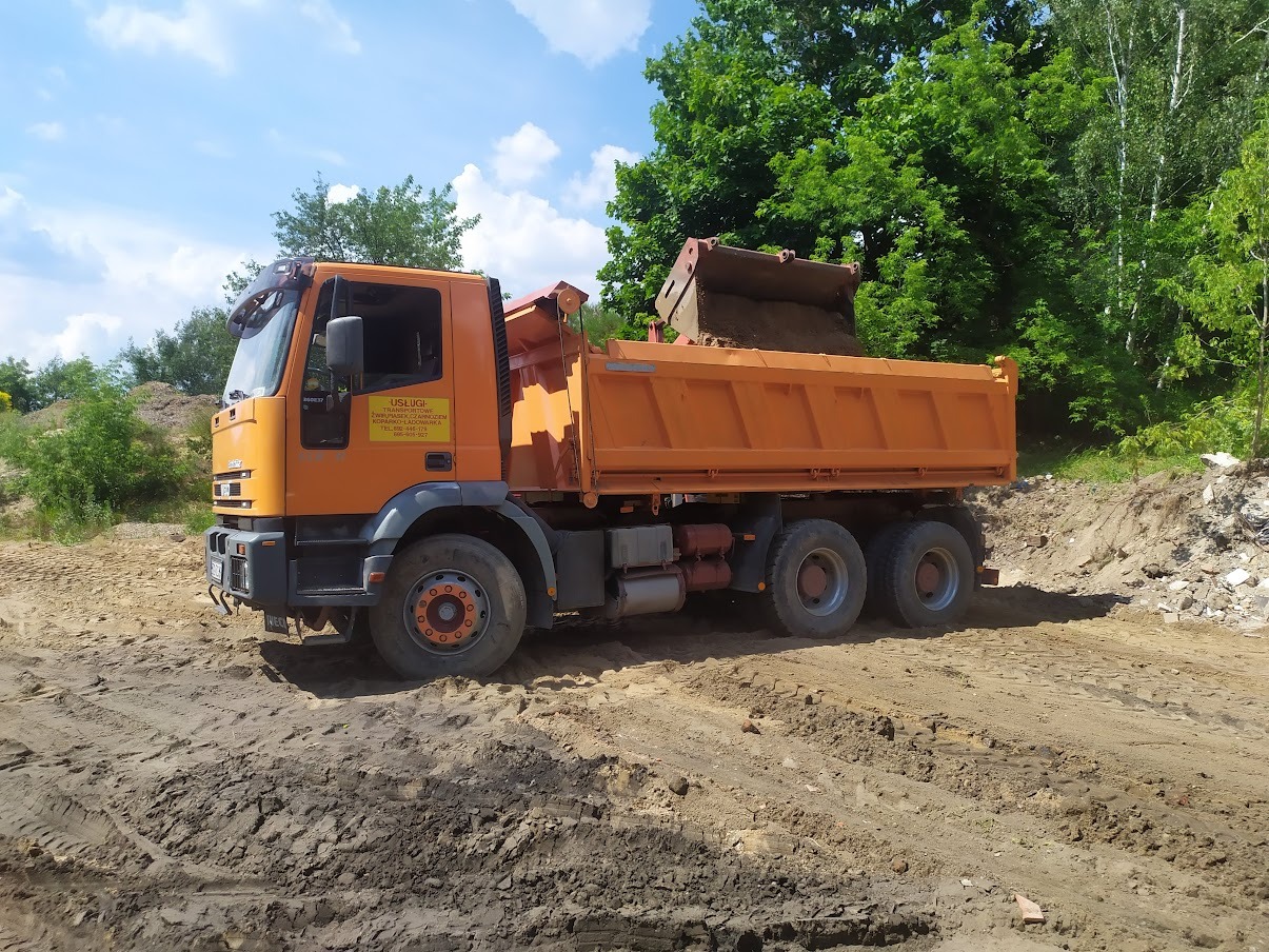 Pomarańczowa wywrotka z ładunkiem ziemi w trakcie pracy na terenie budowy, widoczne ślady opon na grząskim podłożu, zieleń drzew w tle, błękitne niebo z chmurami.