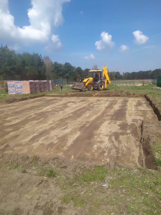 Żółta koparko-ładowarka przygotowuje teren pod budowę, widoczne wykopane obrysy fundamentów oraz palety z cegłami w tle.