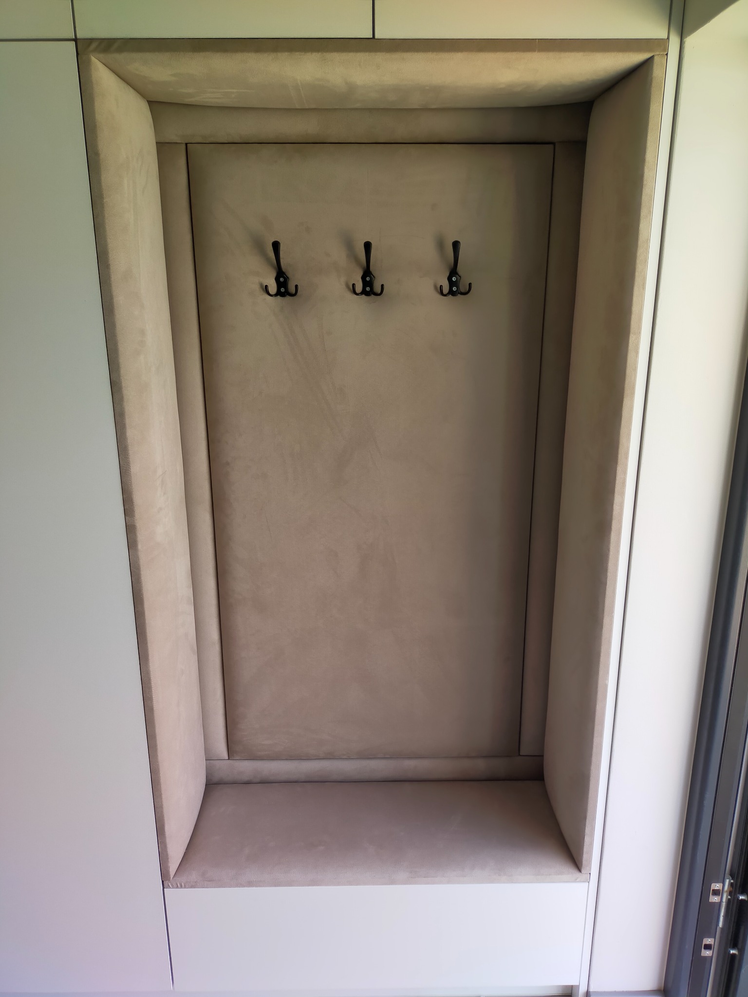 Wnęka z siedziskiem i wieszakami w przedpokoju, obita beżową tapicerką, wkomponowana w białą zabudowę meblową.