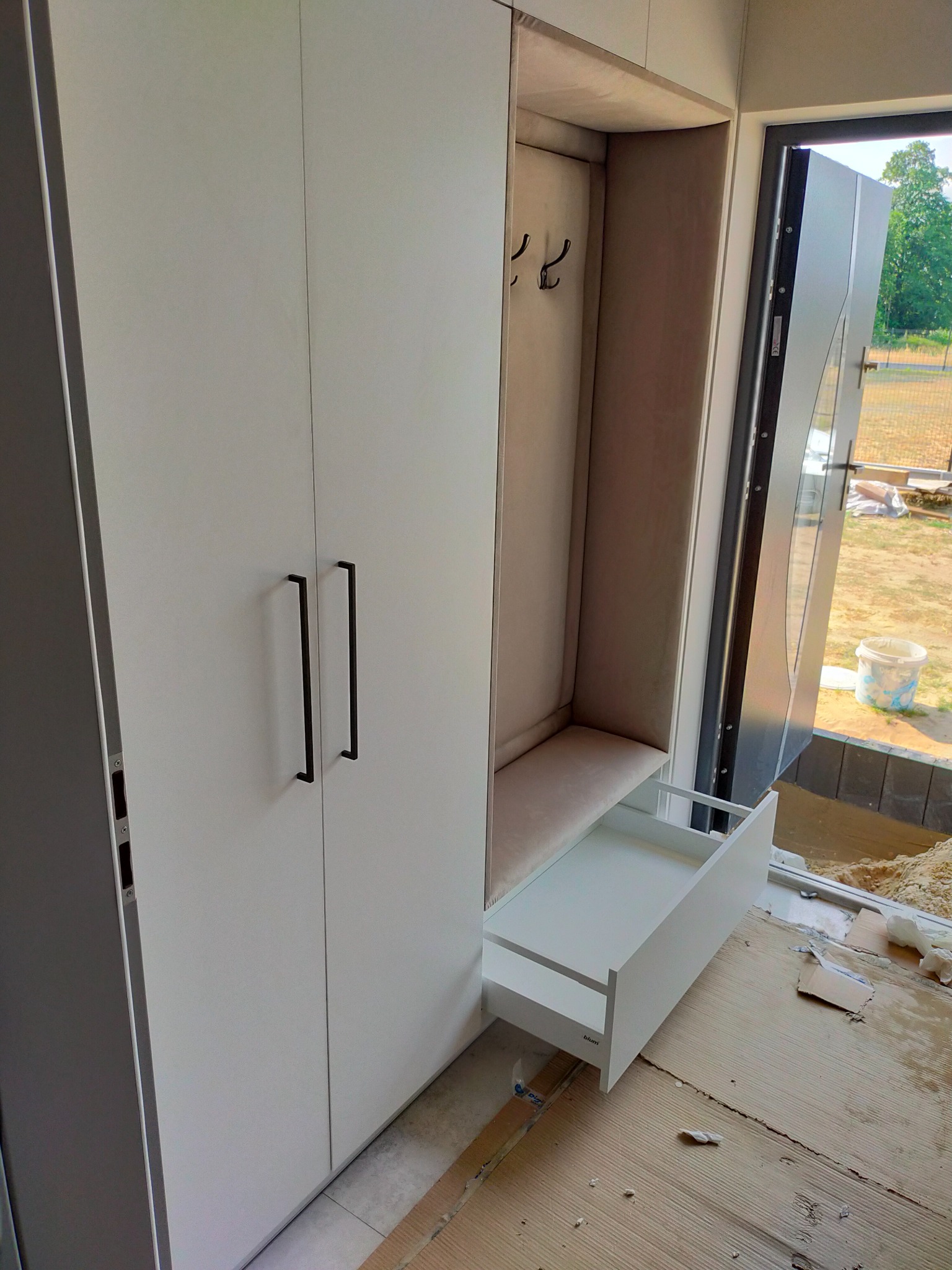 Biała szafa z otwartą wnęką na ubrania i wysuwaną szufladą, widoczna w trakcie montażu w Grodzisku Wielkopolskim, z elementami wykończeniowymi leżącymi na podłodze.