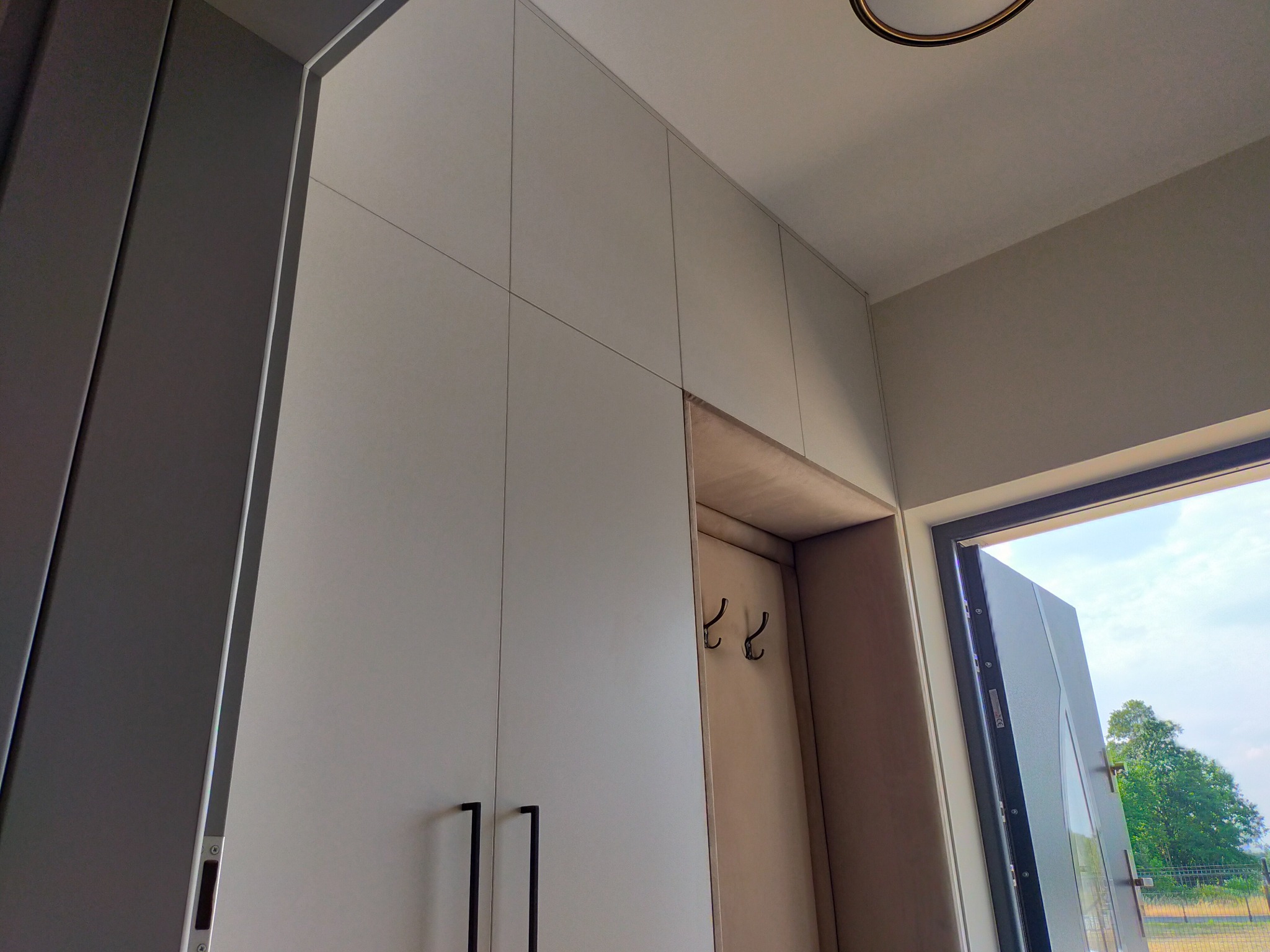 Biała szafa w przedpokoju z czarnymi uchwytami, wnęka z wieszakami i widok na zewnątrz przez otwarte drzwi.