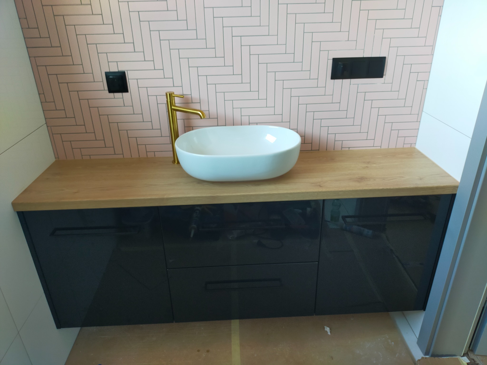 Nowoczesna łazienka z umywalką nablatową, złotą armaturą i szafką z ciemnymi, lakierowanymi frontami pod blatem z jasnego drewna, na tle ściany wyłożonej różową cegiełką ułożoną w jodełkę.