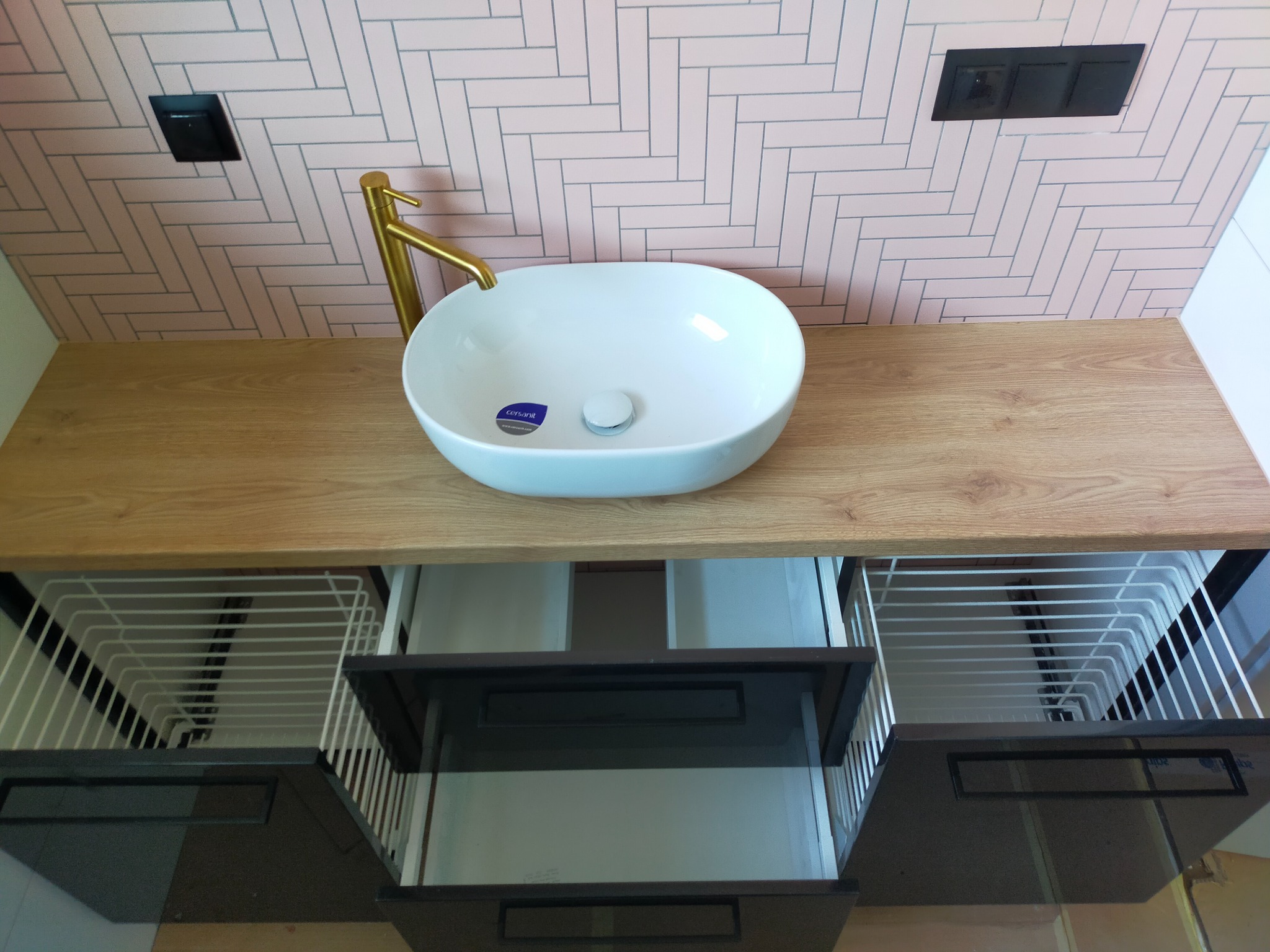 Nowoczesna łazienka z białą umywalką nablatową, złotą baterią i blatem z jasnego drewna, różowe płytki ułożone w jodełkę na ścianie, otwarte szuflady z metalowymi koszami.