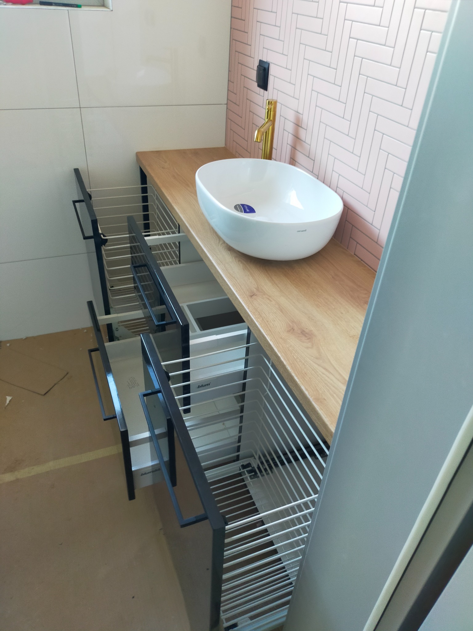 Łazienkowa szafka pod umywalkę z wysuwanymi koszami, drewniany blat i biała umywalka nablatowa na tle różowej ściany w jodełkę, widoczne detale szuflad Blum.