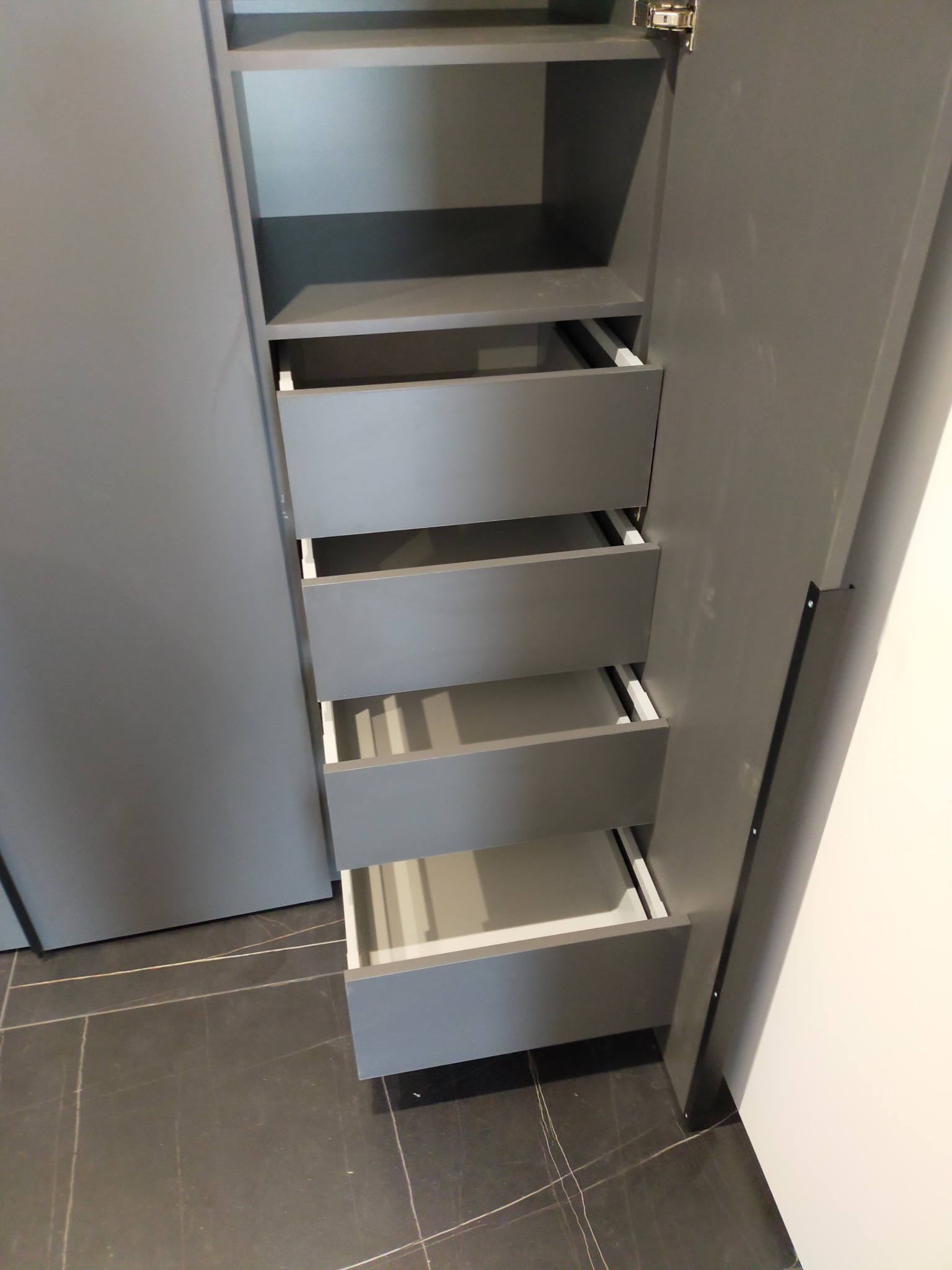 Wnętrze szarej szafy z wysuwanymi szufladami, widoczne półki i system prowadnic, ujęcie z góry na tle ciemnej podłogi.