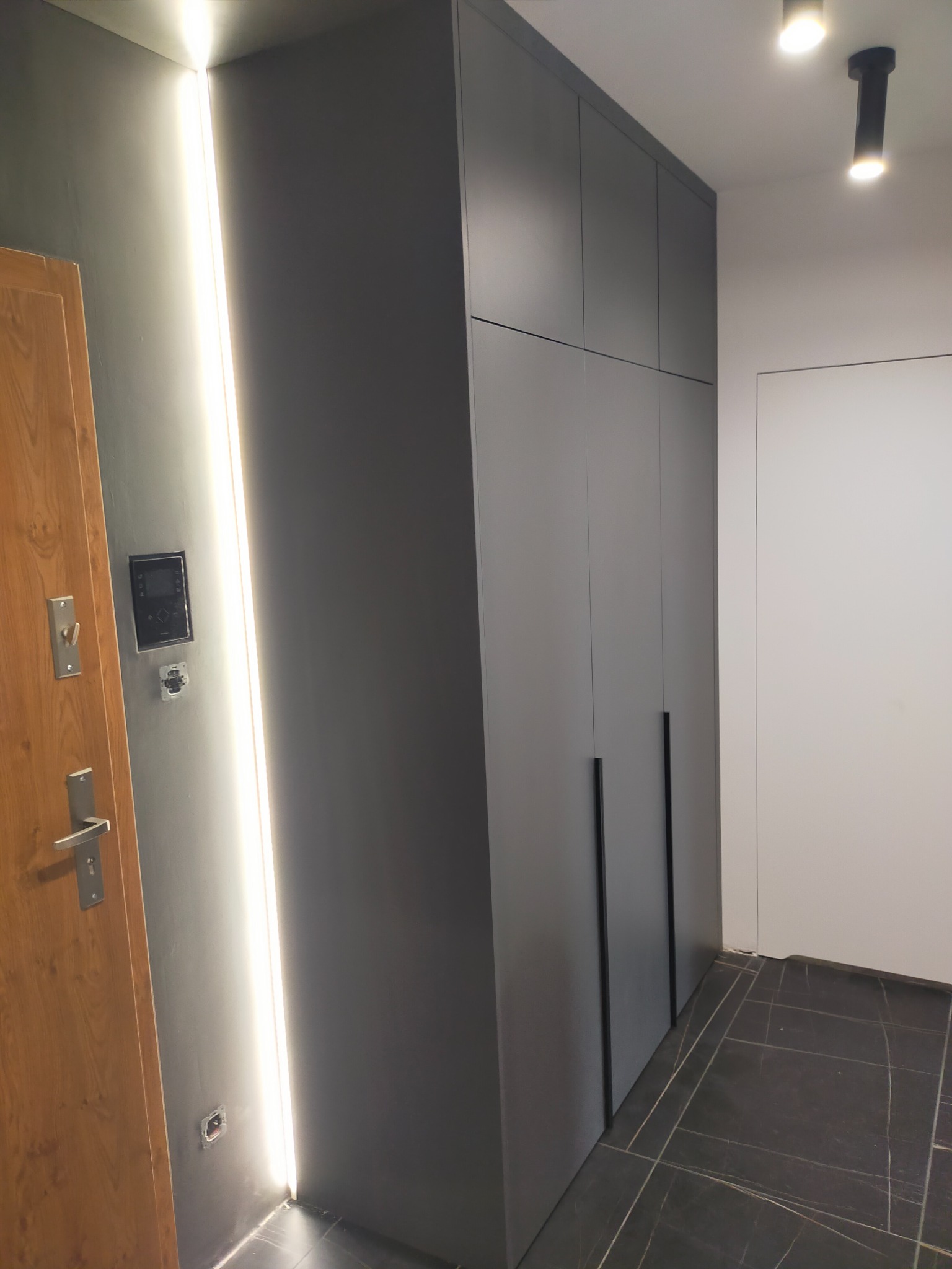 Szara szafa wnękowa z długimi, pionowymi uchwytami, wkomponowana w przedpokój z drewnianymi drzwiami i podświetleniem LED w ścianie.