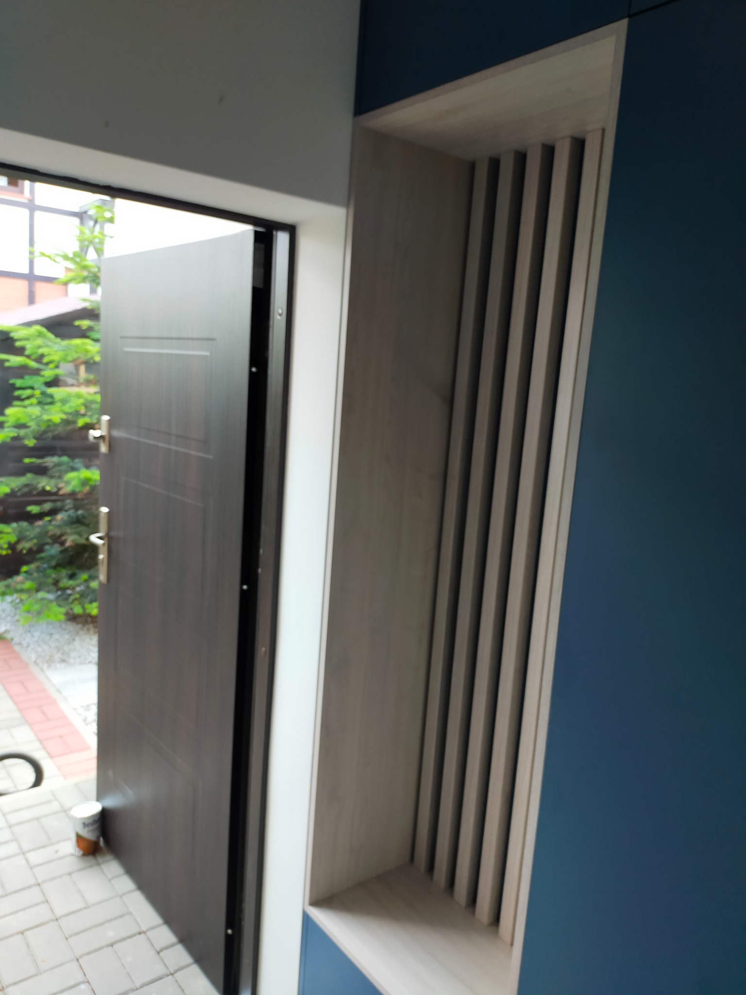 Ciemne, częściowo otwarte drzwi wejściowe z drewnianym panelem dekoracyjnym z pionowych listew po prawej stronie, widok z wnętrza domu na zewnątrz.