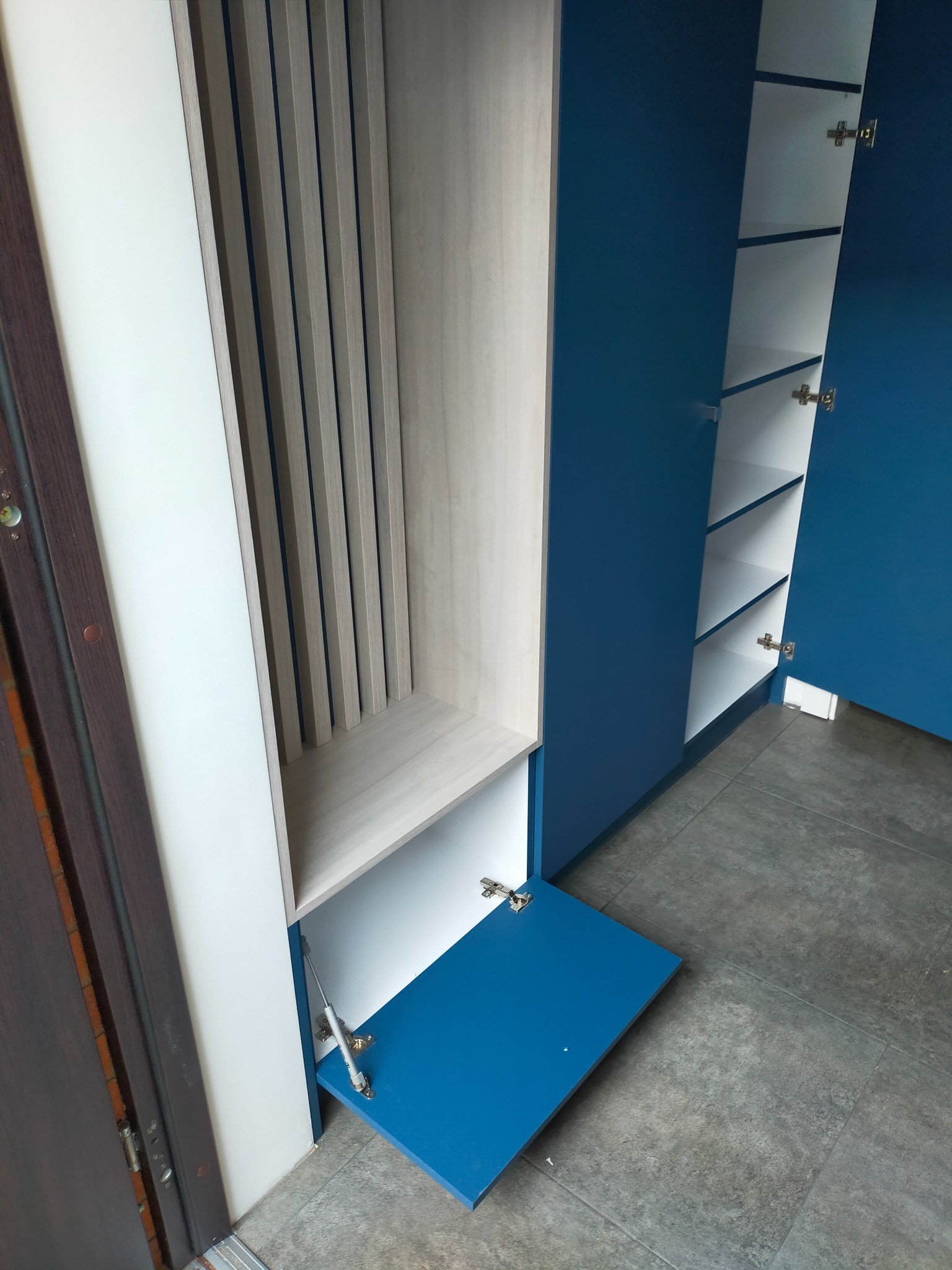 Niebiesko-biała szafa z otwartymi drzwiami i półkami, widoczne metalowe zawiasy i mechanizm podnoszenia klapy, szara podłoga.