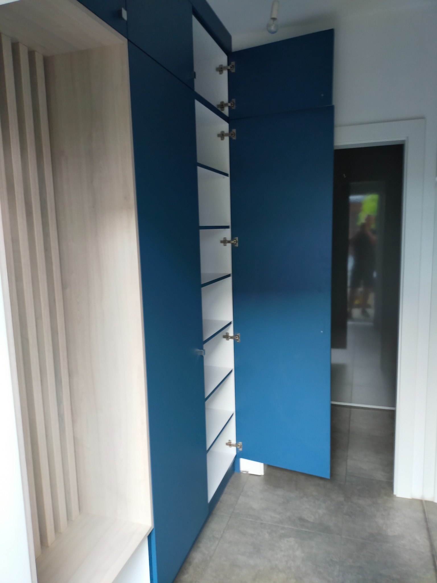 Niebieska szafa z otwartymi drzwiami ukazująca białe półki, obok regał z jasnego drewna z pionowymi listwami.