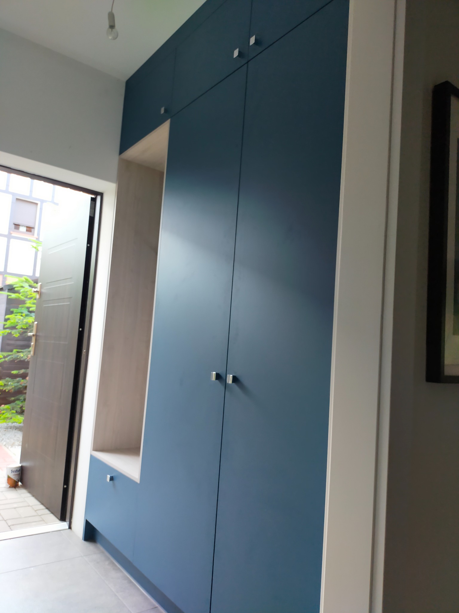 Niebieska szafa wnękowa z jasnym regałem, widoczna przy wejściu do domu z otwartymi drzwiami.