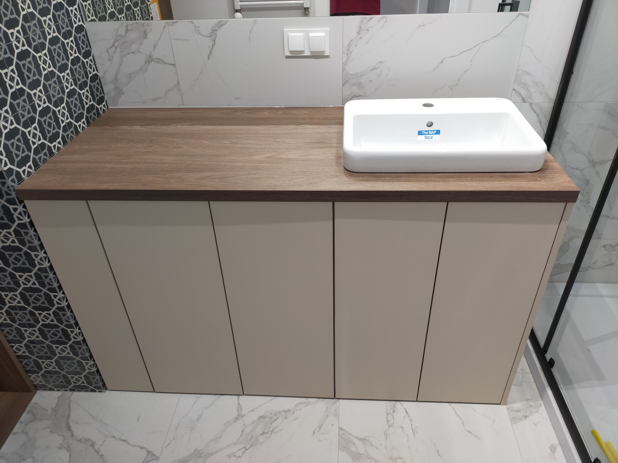 Nowoczesna łazienka z szafką pod umywalkę z drewnianym blatem, białą umywalką Roca Gap i marmurowymi płytkami na ścianach.