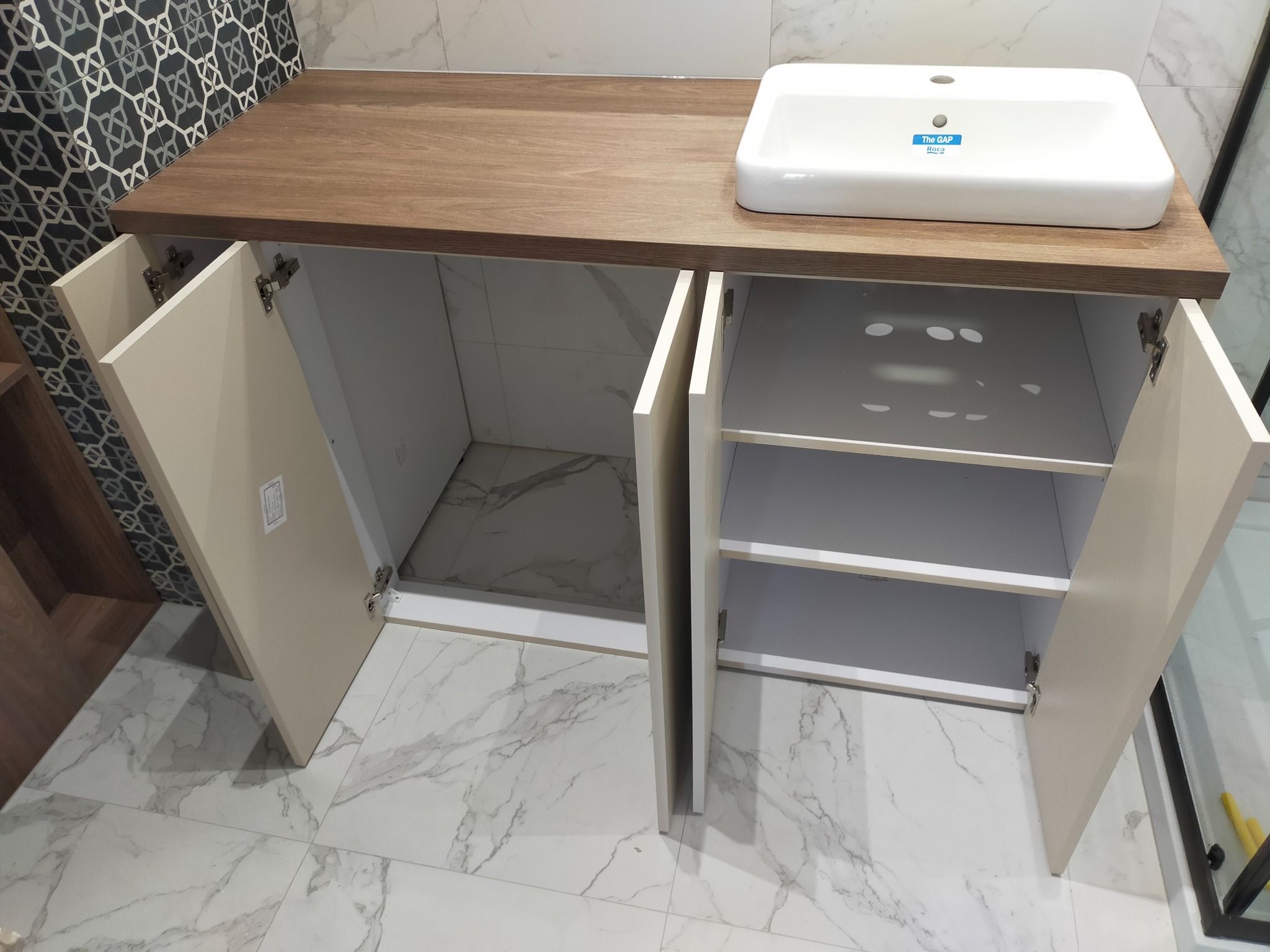 Łazienkowa szafka pod umywalkę z blatem drewnopodobnym, otwarte kremowe fronty szafek, biała umywalka nablatowa Roca, marmurowe płytki na podłodze i ścianie.