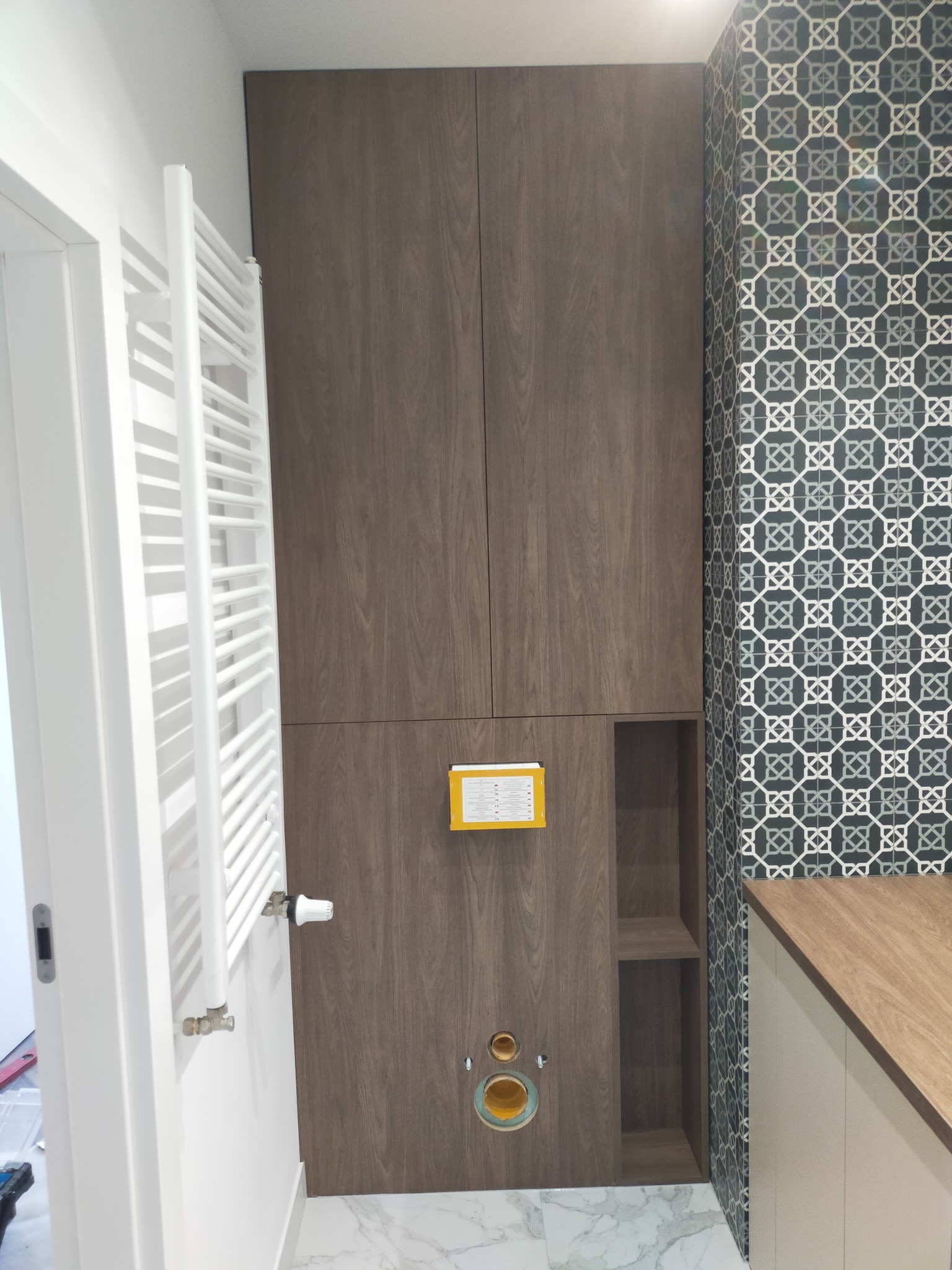 Nowoczesna łazienka z zabudową meblową w kolorze ciemnego drewna, dekoracyjnymi płytkami na ścianie i białym grzejnikiem drabinkowym, widoczne przyłącza kanalizacyjne