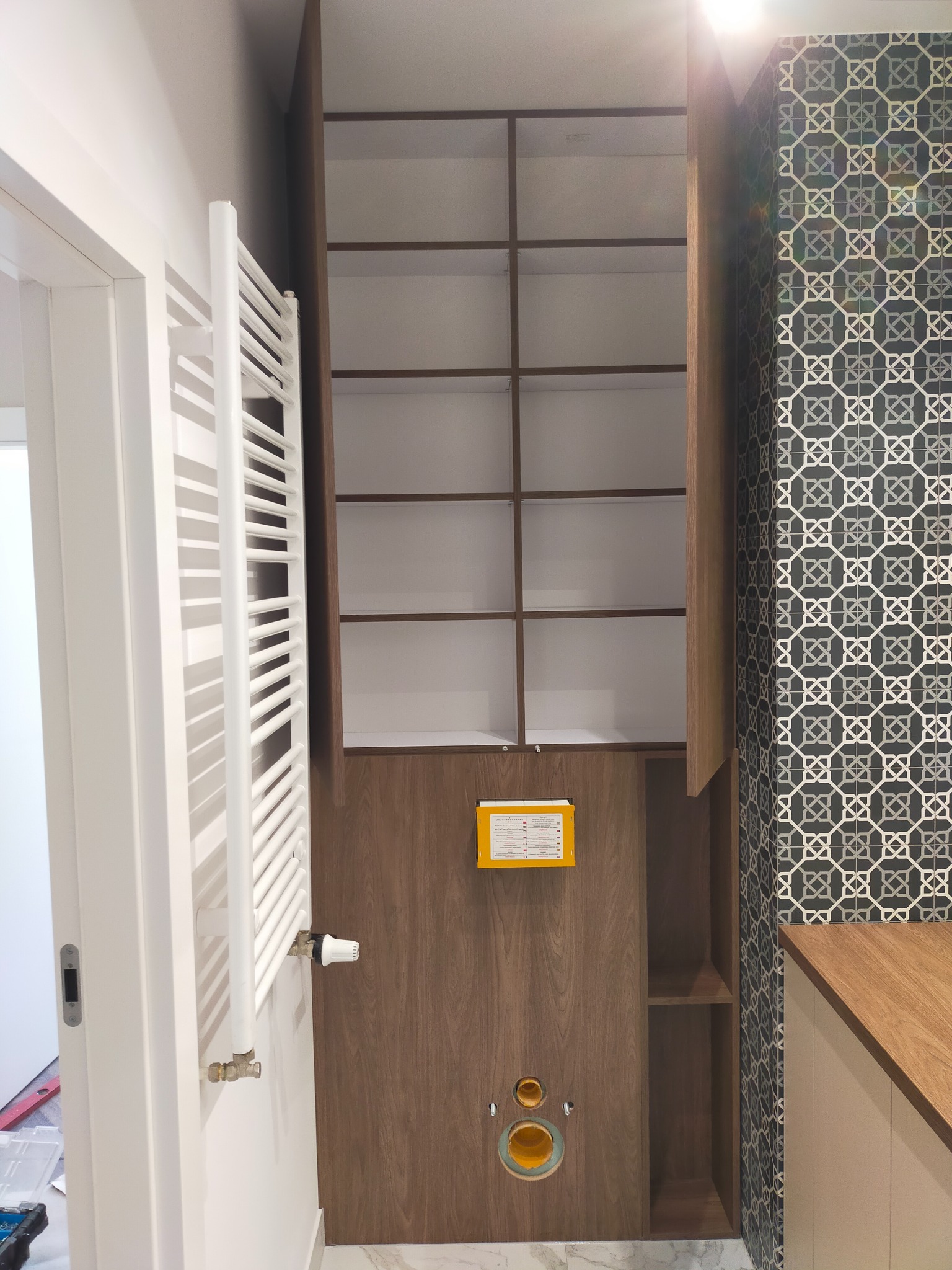 Wnętrze łazienki z drewnianą zabudową szafki wiszącej z otwartymi półkami, dekoracyjną mozaiką na ścianie i białym grzejnikiem drabinkowym.