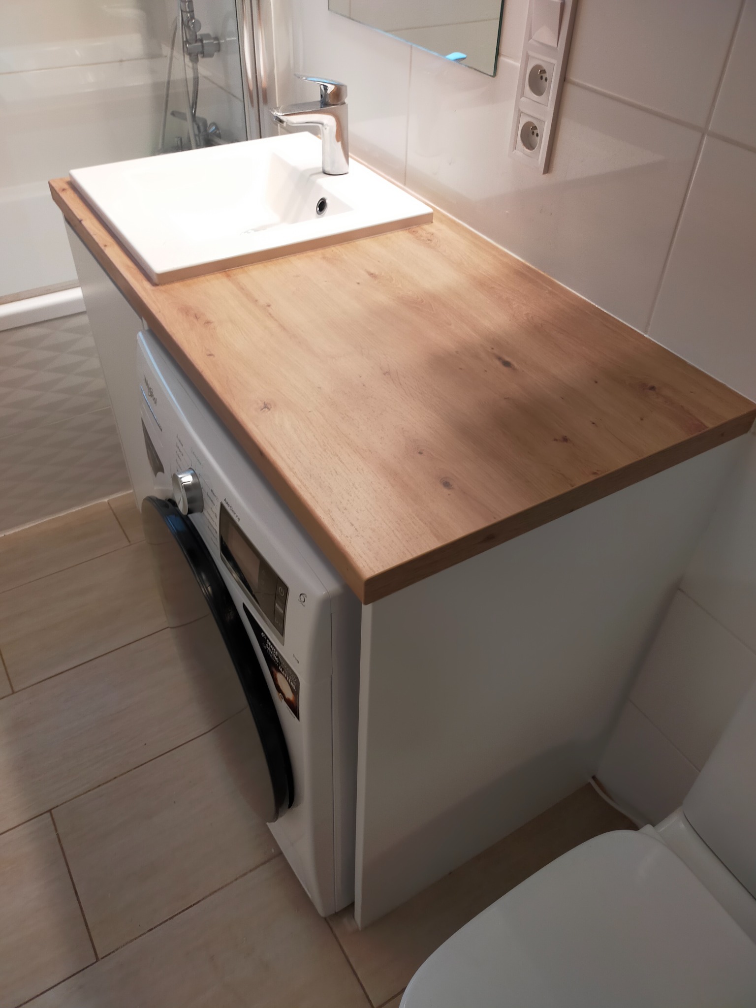 Biała umywalka nablatowa z chromowaną baterią, zamontowana na blacie z jasnego drewna, pod którym znajduje się pralka w zabudowie; łazienka z jasnymi płytkami.