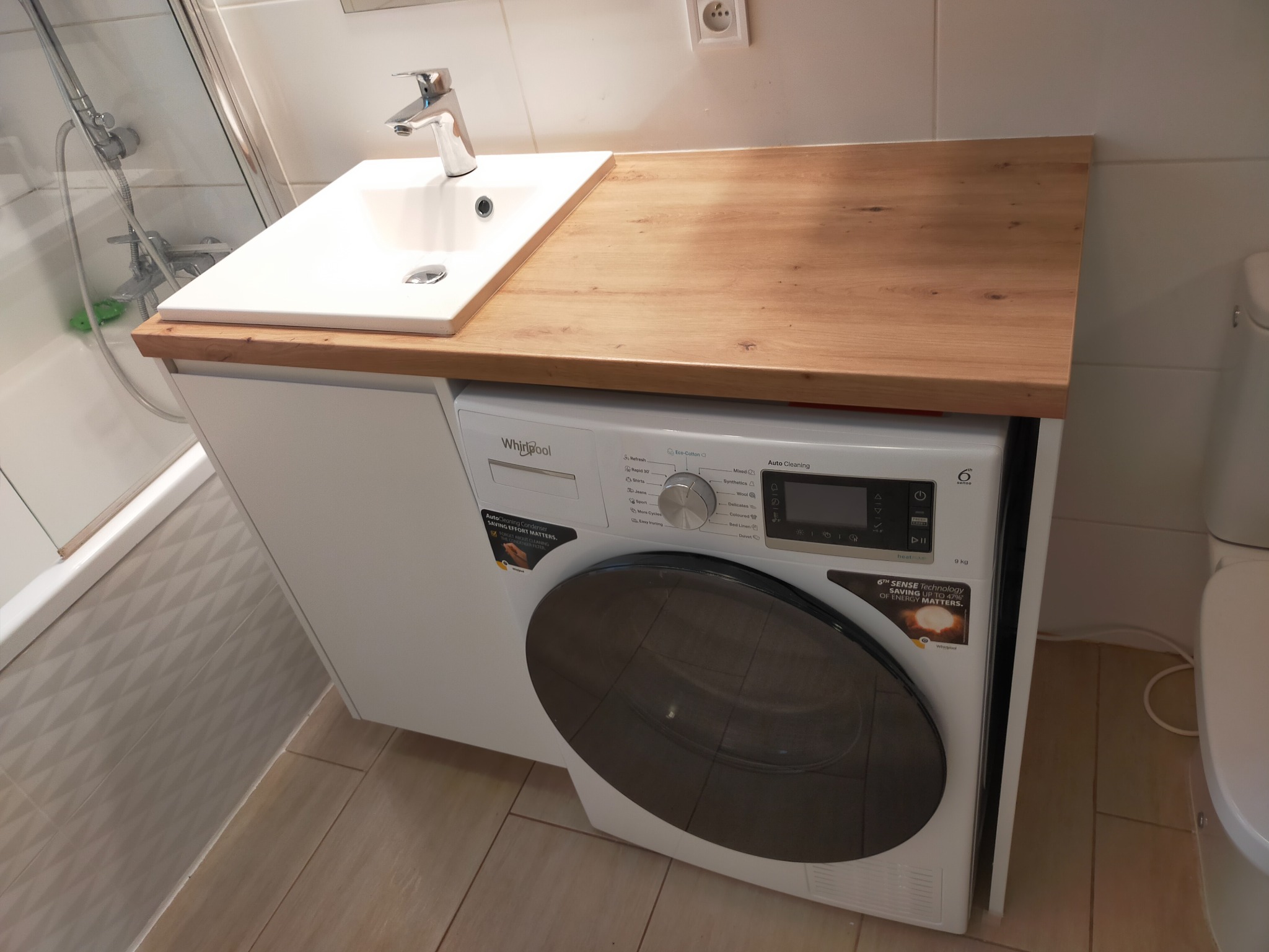 Łazienka z pralką zabudowaną pod drewnianym blatem z umywalką nablatową, widok na baterię, białe szafki i beżowe płytki podłogowe.