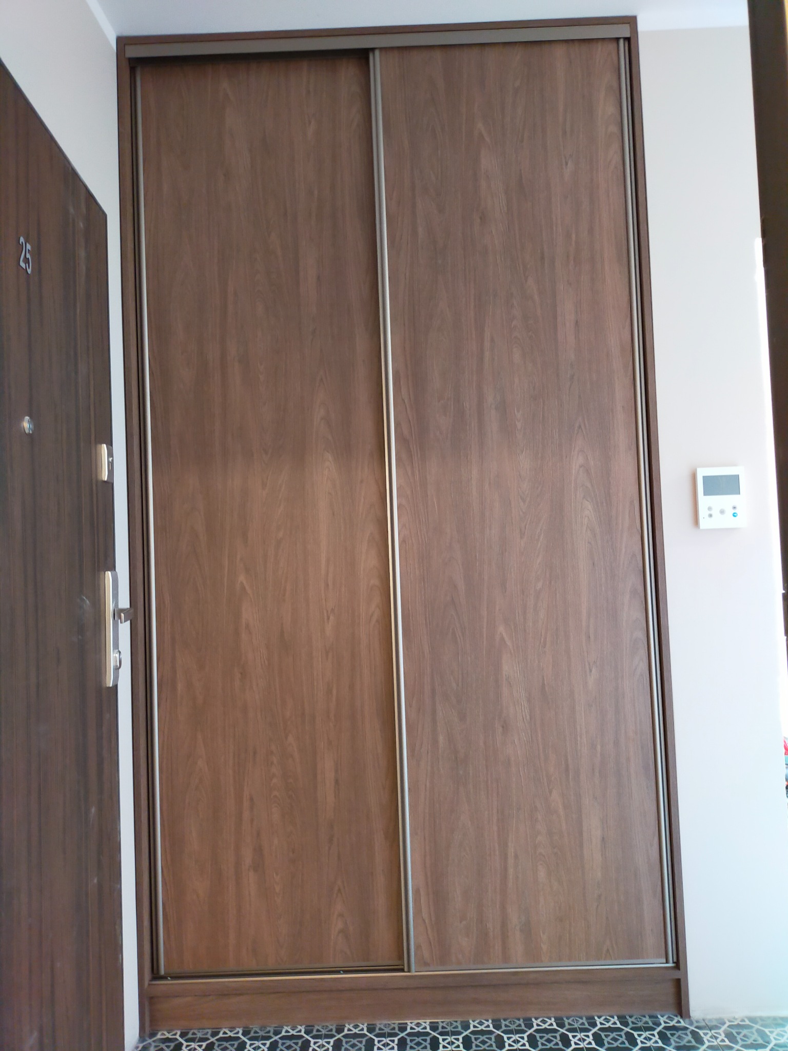 Wnętrze przedpokoju z zabudowaną szafą przesuwną o drewnopodobnym wykończeniu, widoczny panel sterowania na ścianie i drzwi wejściowe z numerem 25.