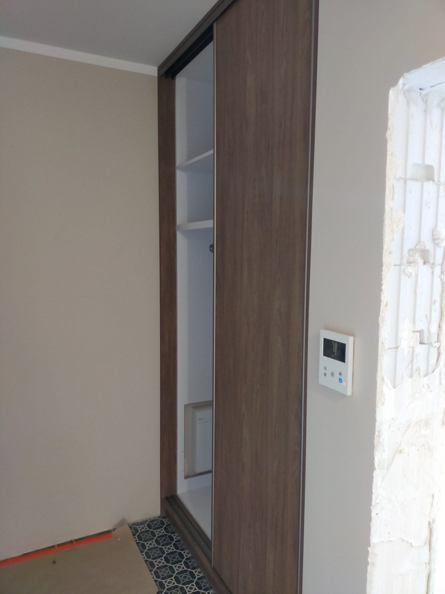 Szafa wnękowa z przesuwnymi drzwiami o drewnianym wykończeniu, widoczne białe półki wewnątrz, obok fragment ściany w trakcie wykończenia i panel sterowania na ścianie.