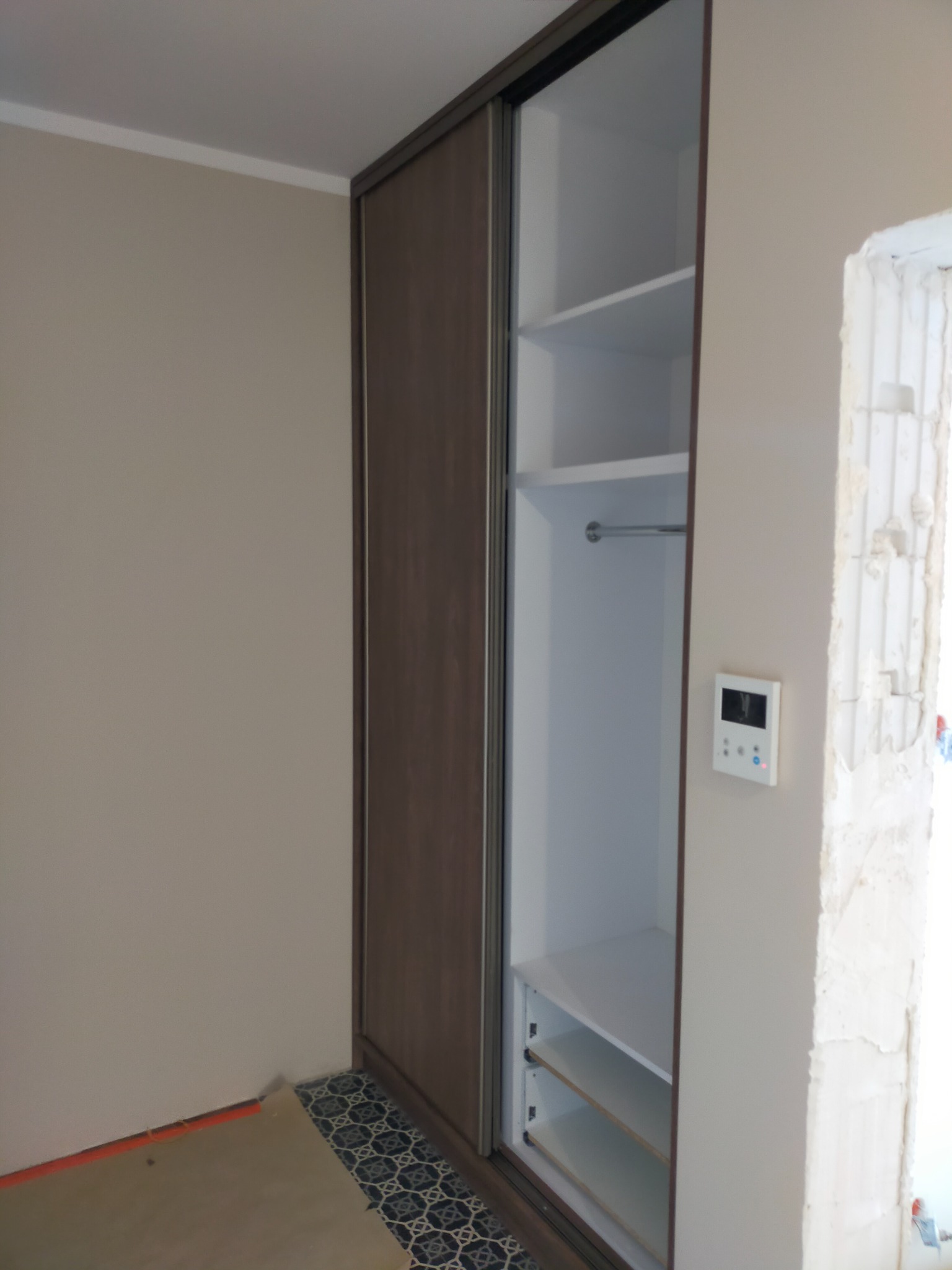 Wnękowa szafa z przesuwnymi drzwiami w kolorze ciemnego drewna, wnętrze białe z półkami i drążkiem, widoczny fragment podłogi z wzorzystymi płytkami, ściana w trakcie wykończenia przy ościeżnicy.