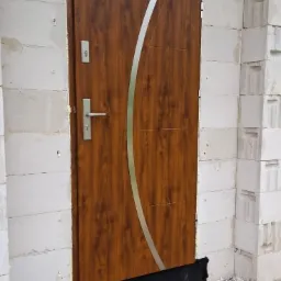 Brązowe drzwi wejściowe z ozdobnym, srebrnym paskiem, zamontowane w surowym murze z betonu komórkowego, widoczne pianka montażowa i czarna izolacja przy progu.