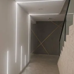 Nowoczesny korytarz z oświetleniem LED wpuszczonym w ścianę, dekoracyjna ściana w tle z geometrycznym wzorem i schody z szklaną balustradą.