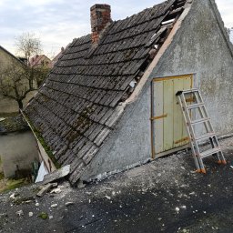 Norbert Konieczny - Zdjęcie dachu z widocznymi uszkodzeniami i mchem, z drabina opartą o ścianę. Fragmenty dachu są usunięte, ukazując konstrukcję. Widoczny komin i otoczenie budynku.