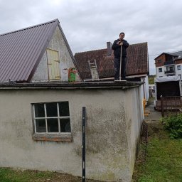 Norbert Konieczny - Pracownik na dachu budynku z nowym, brązowym pokryciem z blachy trapezowej. Widoczna drabina, butla gazowa i poziomica. Praca na wysokościach w pochmurny dzień.