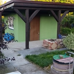 Norbert Konieczny - Zielony budynek gospodarczy z brązowym dachem i drewnianą konstrukcją wsporczą. Widoczny fragment ogrodu z elementami dekoracyjnymi i roślinnością.