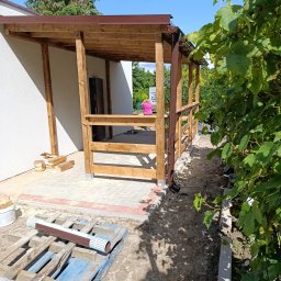 Norbert Konieczny - Drewniana, zadaszona weranda w trakcie budowy. Widoczne elementy konstrukcyjne, brązowe rynny, paleta z materiałami i fragment ogrodu z winoroślą.