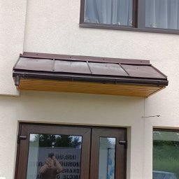 Norbert Konieczny - Brązowy daszek nad wejściem do budynku z brązowymi oknami. Widoczne odbicie osoby robiącej zdjęcie w szybie drzwi.