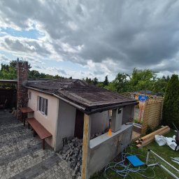 Norbert Konieczny - Przygotowania do wymiany dachu na budynku gospodarczym z kominem. Stare pokrycie dachowe, obok kostka brukowa i materiały budowlane.