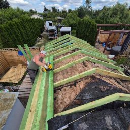 Norbert Konieczny - Prace dekarskie: mężczyzna w trakcie montażu drewnianej konstrukcji dachu, widoczne ocieplenie i narzędzia. W tle zieleń drzew i zabudowania.