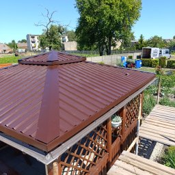 Norbert Konieczny - Nowy, brązowy dach z blachy trapezowej na altanie ogrodowej z drewnianą konstrukcją kratową. Widoczne rynny i fragment ogrodu w tle.