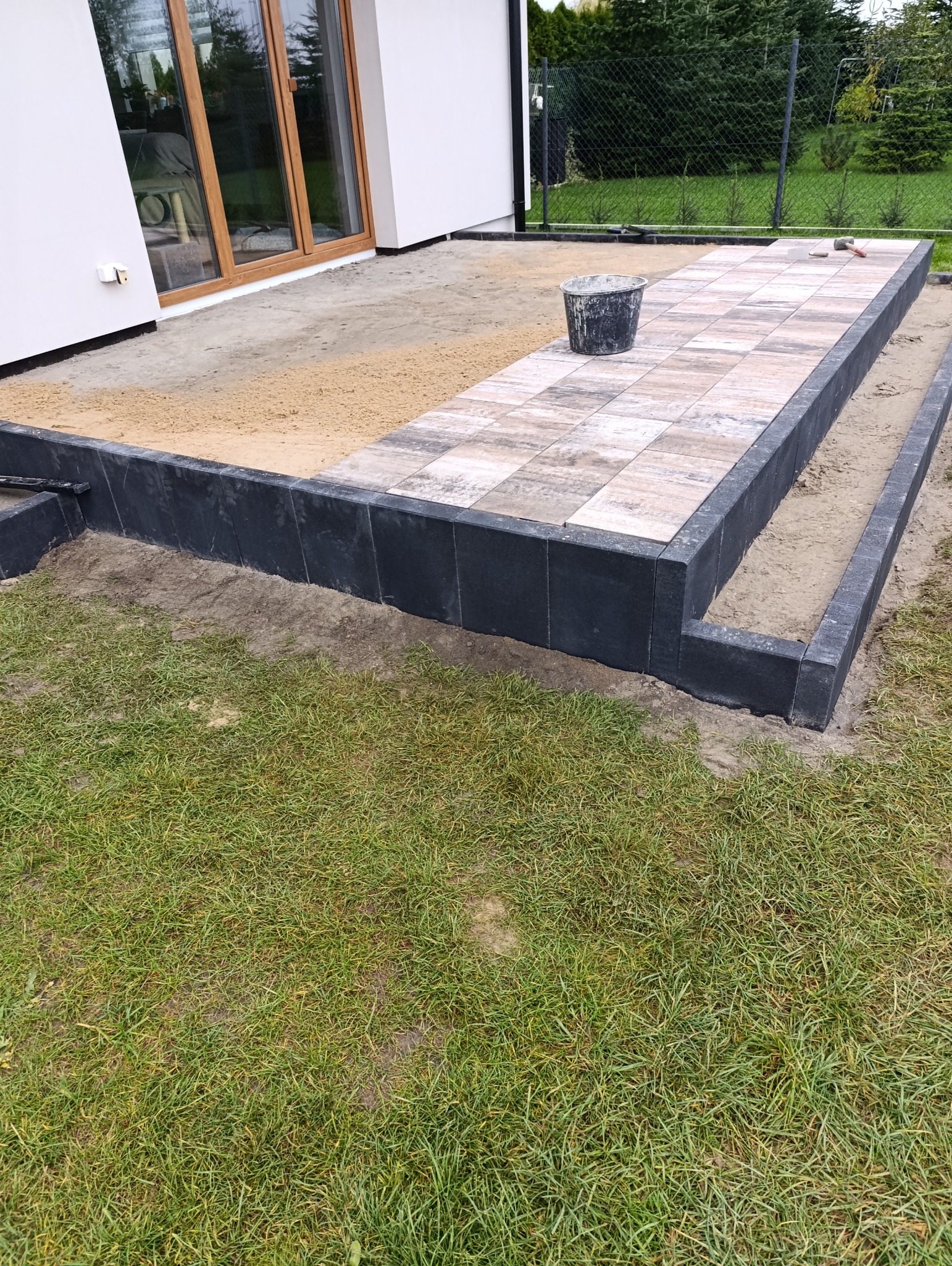 Układanie kostki brukowej na tarasie domu jednorodzinnego, widok z poziomu trawnika. Częściowo ułożona kostka, czarne obrzeża, wiadro na kostce.