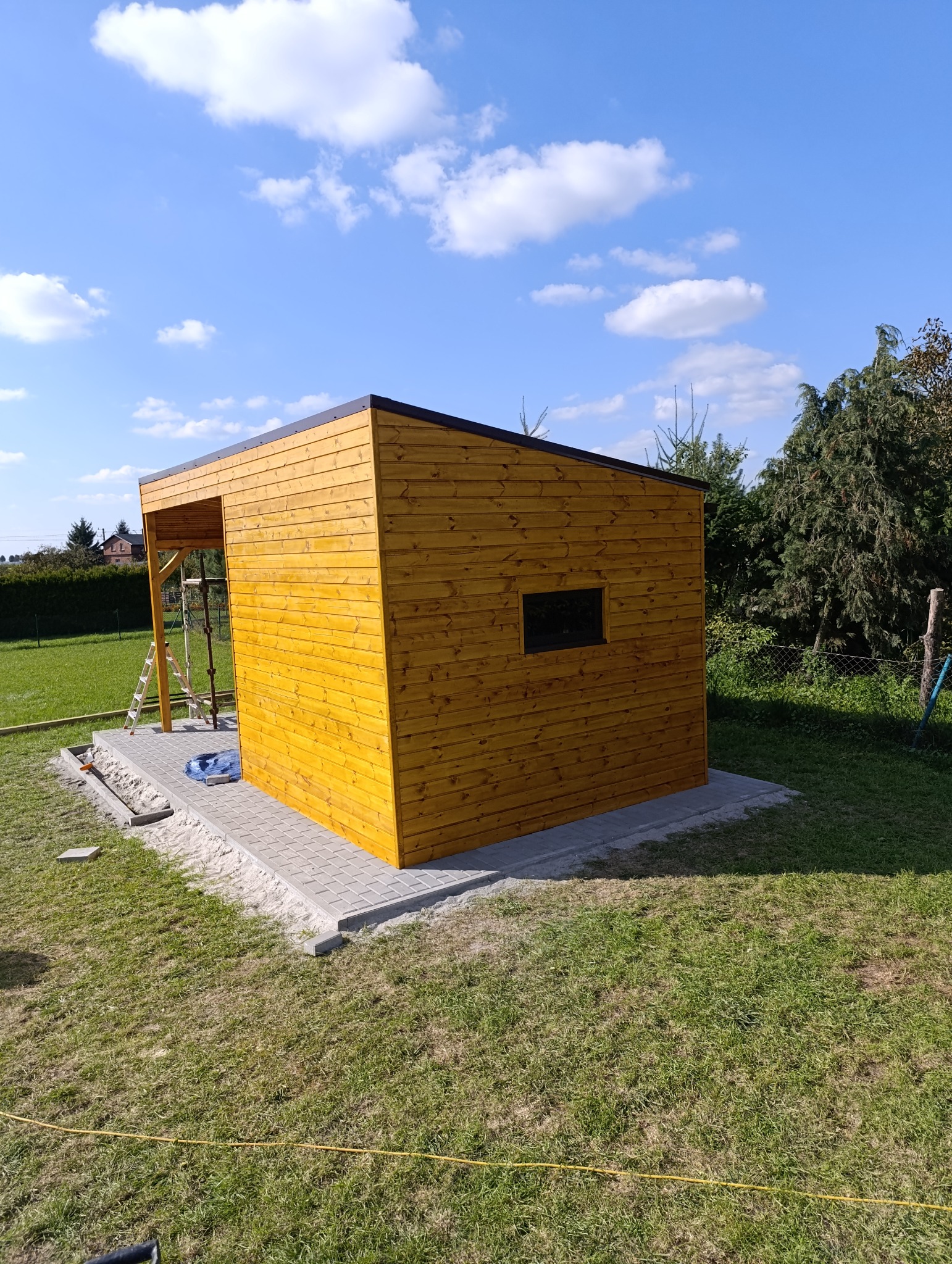 Nowoczesna, drewniana altana ogrodowa z płaskim dachem, świeżo po montażu, na tle zielonego trawnika i błękitnego nieba z chmurami. Widoczna drabina i kostka brukowa.