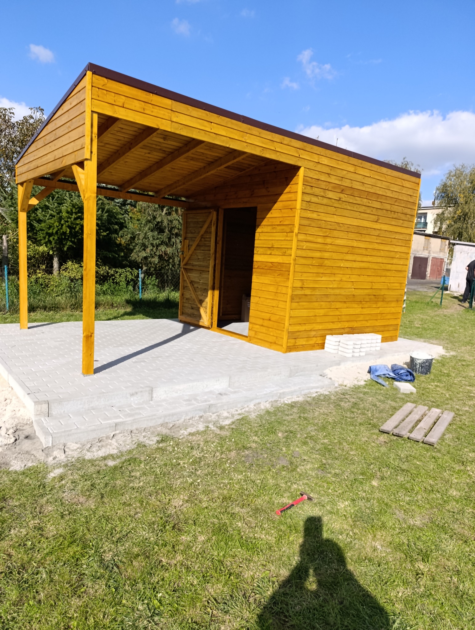 Drewniana altana z brązowym dachem, częściowo otwarta, z betonową podstawą. Widoczne narzędzia i materiały budowlane. Słoneczny dzień, błękitne niebo.