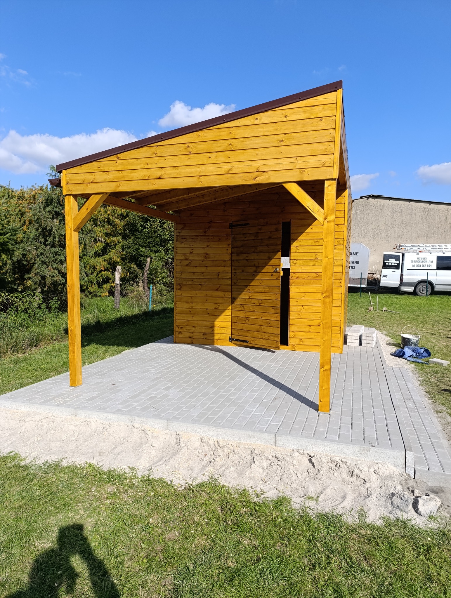 Drewniana altana z brązowym dachem, świeżo po montażu, na tle zieleni i błękitnego nieba. Widoczny fragment ułożonej kostki brukowej i cień osoby robiącej zdjęcie.