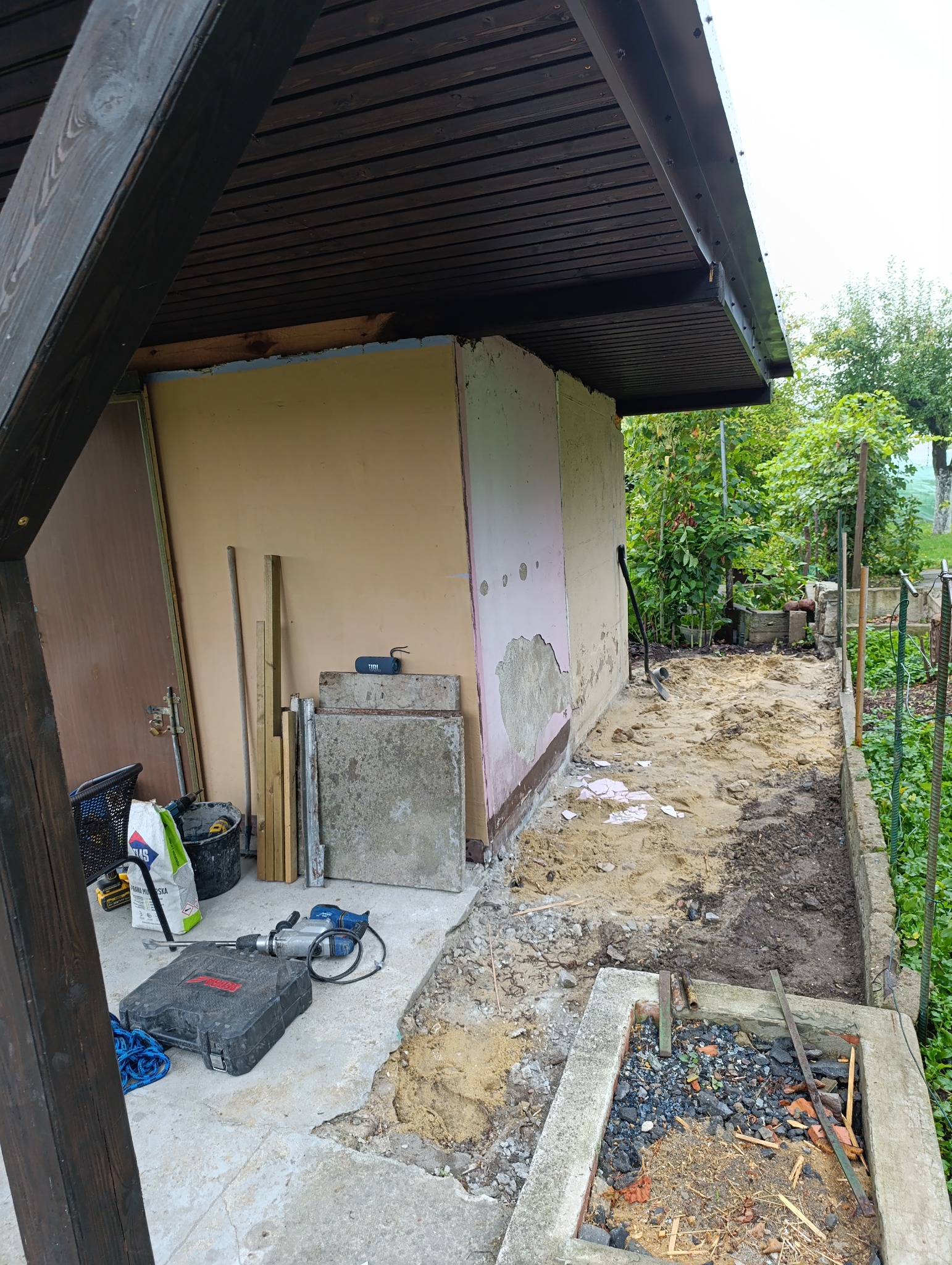 Przygotowania do ocieplenia ścian zewnętrznych budynku gospodarczego. Widoczne narzędzia, materiały budowlane i częściowo usunięty tynk. Prace w toku, teren wokół rozkopany.