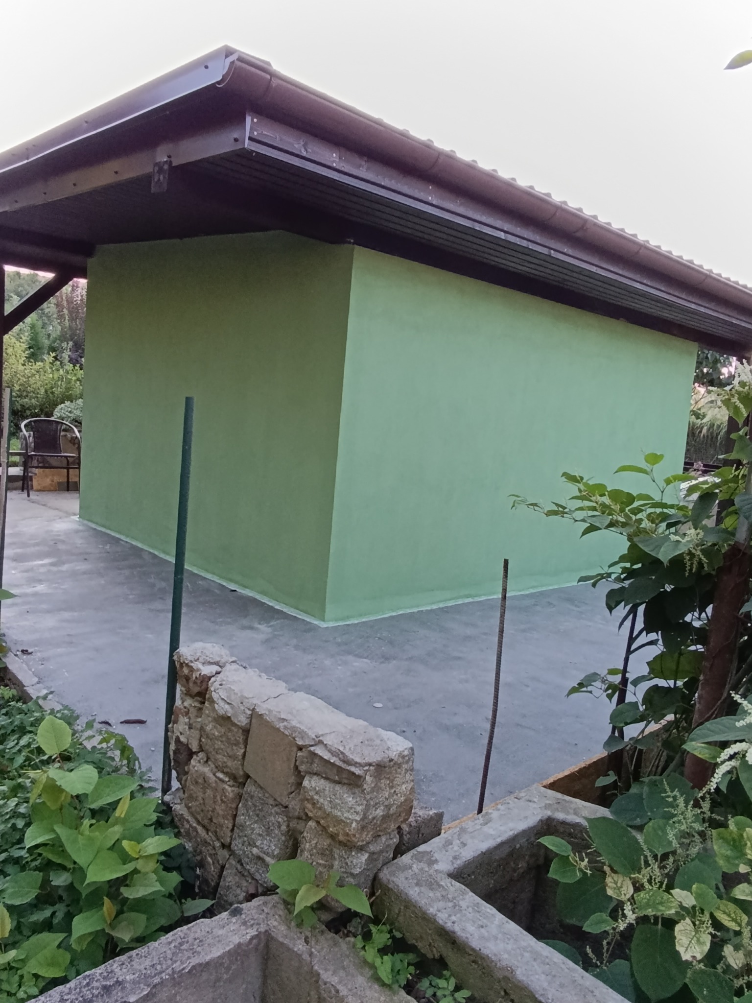 Świeżo pomalowany na zielono budynek gospodarczy z brązowym dachem, widoczny z poziomu ogrodu. Kamienna rabata w pierwszym planie.