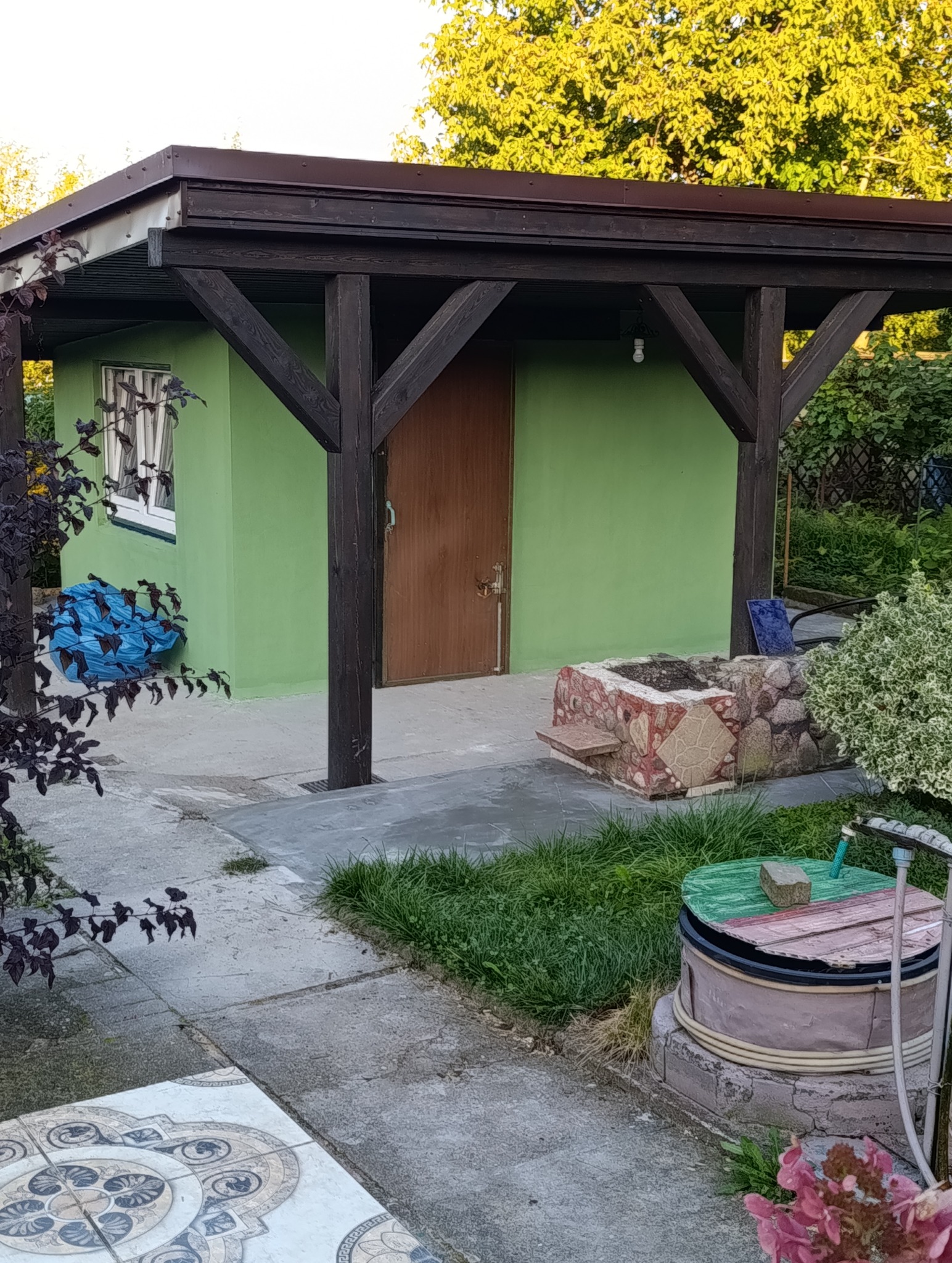 Zielony budynek gospodarczy z brązowym dachem i drewnianą konstrukcją wsporczą. Widoczny fragment ogrodu z elementami dekoracyjnymi i roślinnością.