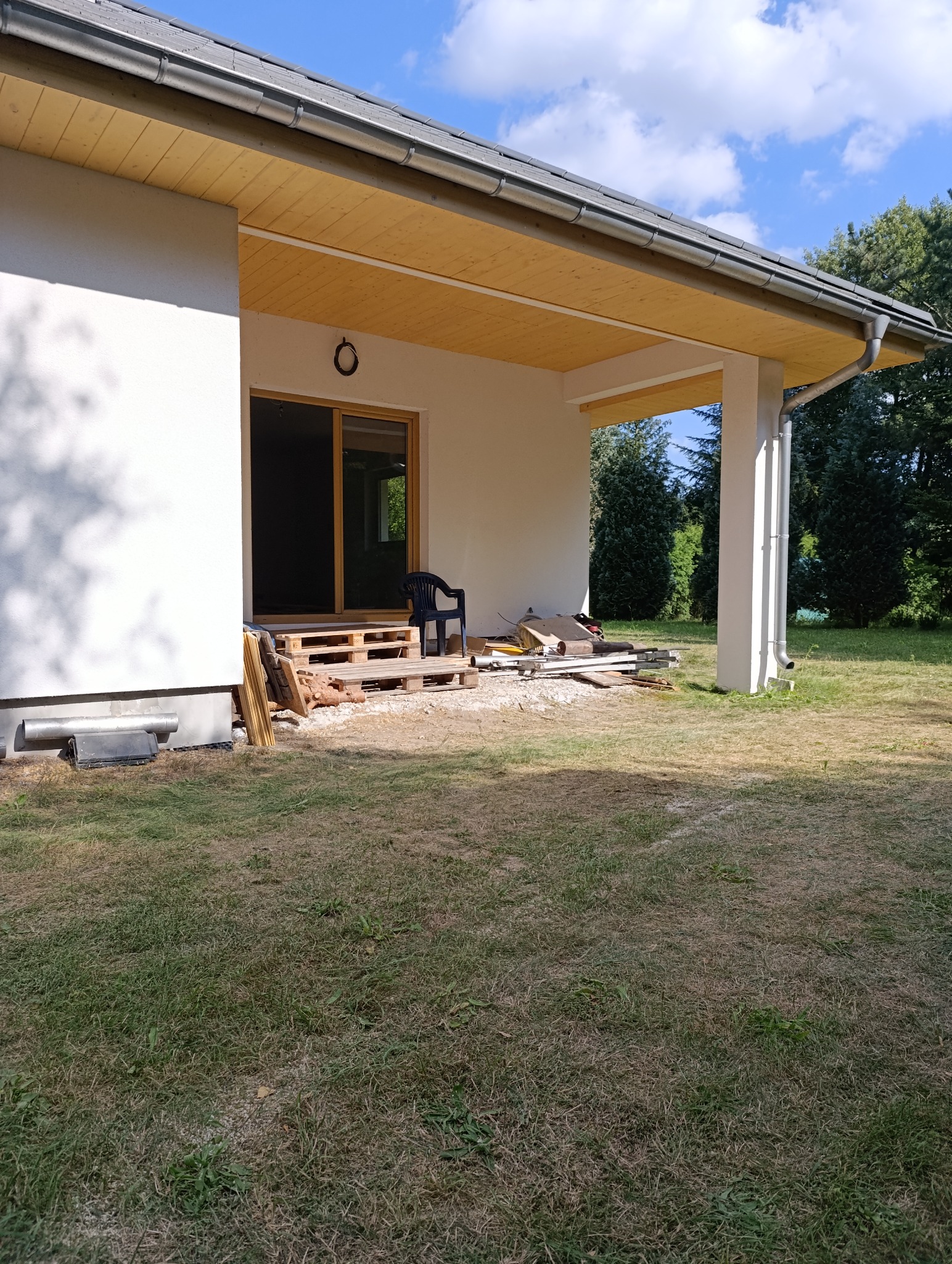 Dom w trakcie budowy z widocznym zadaszeniem, otwartymi drzwiami balkonowymi i materiałami budowlanymi na zewnątrz. Widoczne elementy dachu i ogólny stan prac.