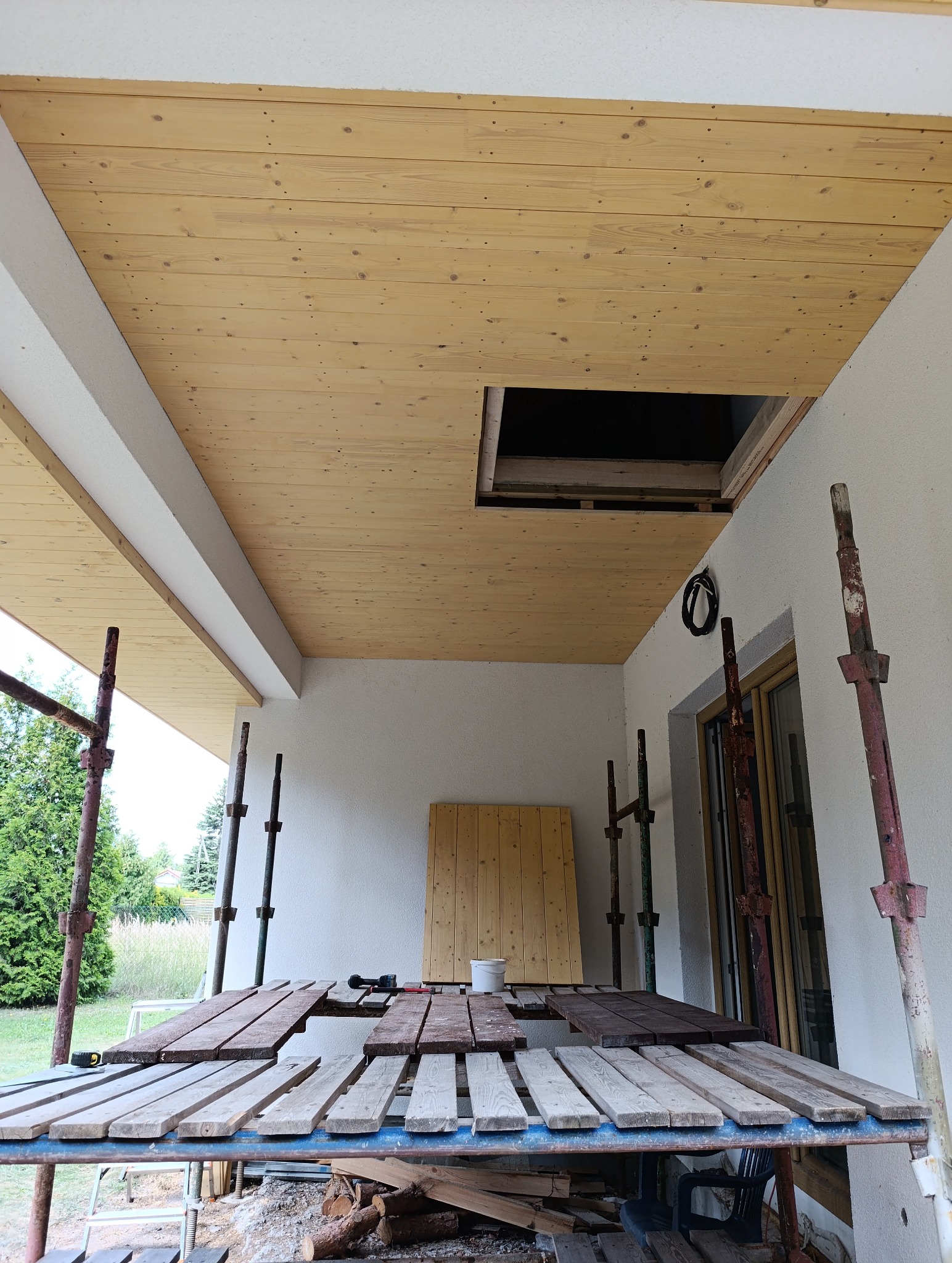 Montaż drewnianej boazerii sufitowej na zewnątrz budynku, widoczne rusztowania i przygotowane deski. Otwór w suficie. Prace wykończeniowe na zewnątrz.